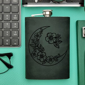 Pode incluir: Uma garrafa de couro preto com uma tampa de prata e um design de lua floral gravado na frente.
