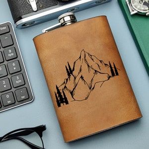 Pode incluir: Uma garrafa de couro marrom com um design de montanha e árvores. A garrafa tem uma tampa prateada e é um ótimo presente para entusiastas de atividades ao ar livre.
