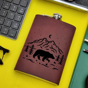 May include: A brown leather flask with a black silhouette of a bear walking through a mountain range with a crescent moon in the sky.