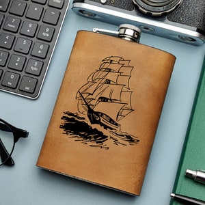 Könnte beinhalten: Eine braune Lederflasche mit einem schwarzen, gravierten Bild eines Segelschiffs mit geschwollenen Segeln. Die Flasche befindet sich auf einer blauen Oberfläche mit einer Tastatur, einer Kamera und einer Uhr im Hintergrund.