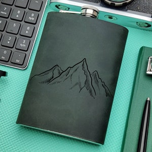 Pode incluir: Uma garrafa de couro verde com uma tampa de prata e um design de cordilheira. A garrafa está sobre uma superfície turquesa com um teclado, uma câmera, um caderno, um relógio e uma caneta.