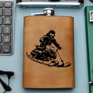 Puede incluir: Una petaca de cuero marrón con una tapa plateada y una silueta negra de una persona montando una moto de nieve.