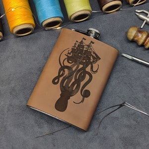 Könnte beinhalten: Eine braune Lederflasche mit silbernem Verschluss und einem gravierten Design eines Kraken, der ein Schiff hält. Die Flasche liegt auf einer grauen Oberfläche mit verschiedenen Werkzeugen und Materialien.