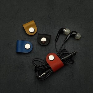 Könnte beinhalten: Vier Leder-Kabelorganizer in Blau, Rot, Schwarz und Gelb mit silbernen Metallverschlüssen. Ein Paar schwarze Ohrhörer sind um den roten Kabelorganizer gewickelt.