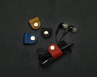 May include: Four leather cord organizers in blue, red, black, and yellow with silver metal clasps. A pair of black earbuds are wrapped around the red cord organizer.