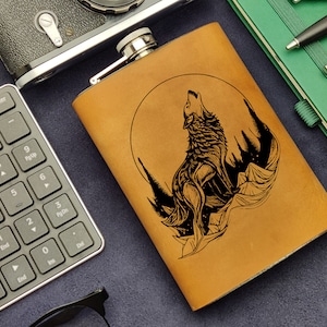 Pode incluir: Uma garrafa de couro marrom com um desenho de lobo preto uivando para a lua. A garrafa está sobre uma superfície azul escura com um teclado, óculos e um caderno verde.