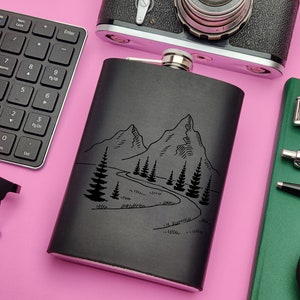 Pode incluir: Frasco de metal preto com um design de montanha e árvores. O frasco está sobre uma superfície rosa.