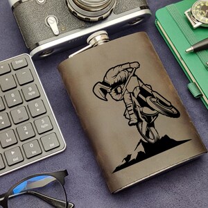 May include: A brown leather flask with a silver top. The flask has a black and white graphic of a motocross rider performing a stunt.