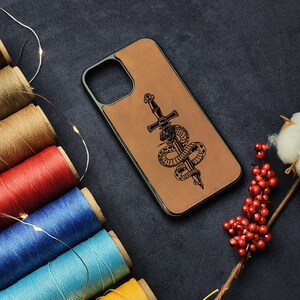 Puede incluir: Una funda de teléfono de cuero marrón con una ilustración en blanco y negro de una serpiente enroscada alrededor de una espada. La funda tiene un recorte para la cámara del teléfono.
