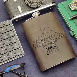 May include: A brown leather flask with a design of a camper van in a forest setting. The flask has the text "TRAVEL more" on it.