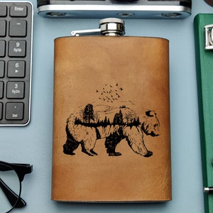 Pode incluir: Uma garrafa de couro marrom com um desenho gravado em preto de um urso com uma floresta dentro. O urso está caminhando para a direita. Há pássaros voando acima do urso.
