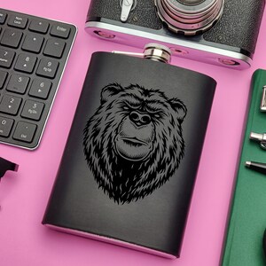 May include: Black metal flask with a black bear head design. The flask is on a pink surface.