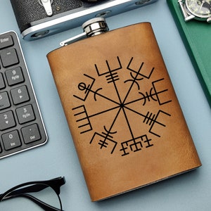 Puede incluir: Una petaca rectangular de cuero marrón con tapa plateada. Presenta un intrincado símbolo de brújula vikinga en negro. La petaca está sobre una superficie azul claro, junto a un teclado, un reloj y un cuaderno.