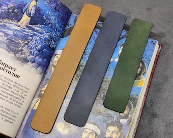 May include: Four leather bookmarks in different colors: brown, dark blue, dark green, and light green. The bookmarks are laying on top of an open book.