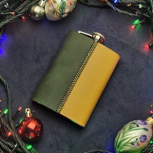 May include: A green and gold leather flask with a zipper detail, surrounded by a string of Christmas lights and ornaments.