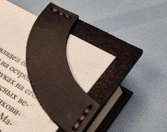 May include: A brown leather bookmark with stitching detail. The bookmark is curved and has a pointed end. It is placed on a book.