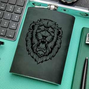 Puede incluir: Una petaca de metal negro con un gráfico de un león rugiente. La petaca está sobre una superficie turquesa.