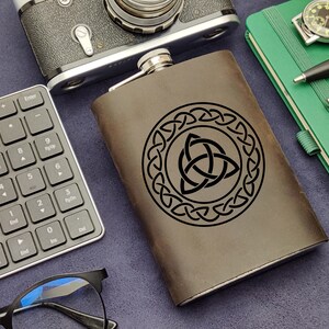 Puede incluir: Una petaca de cuero marrón con un diseño de nudo celta grabado en negro. La petaca está sobre una superficie azul con un teclado, gafas y una libreta.