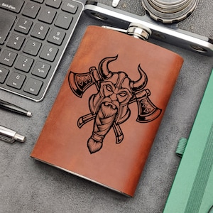 Puede incluir: Una petaca de cuero marrón con una tapa de metal y un diseño grabado en negro de un guerrero vikingo con hachas.
