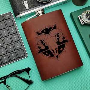 Puede incluir: Una petaca de cuero marrón con un diseño grabado en blanco y negro de una calavera con cuernos dentro de una estrella de cinco puntas. La petaca está sobre una superficie turquesa.