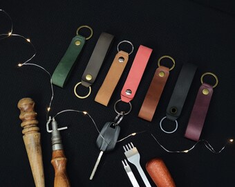 May include: Assortment of eight leather keychains in various colors, including green, brown, tan, pink, black, and burgundy. The keychains are arranged in a row on a black surface.