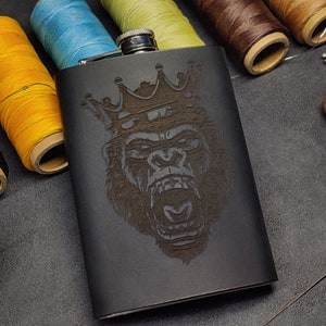 May include: Black leather flask with a silver lid and a laser-engraved design of a gorilla wearing a crown.
