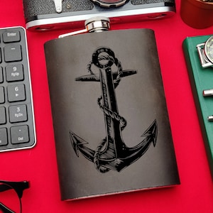May include: A black leather flask with a silver top and a black and white anchor design. The flask is on a red surface.