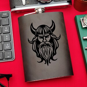Puede incluir: Una petaca de cuero negro con un diseño grabado con láser de un guerrero vikingo con barba y casco.