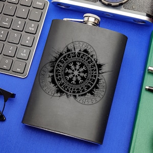 Puede incluir: Una petaca de metal negro con una tapa plateada y un diseño de un símbolo circular con runas alrededor. El símbolo es una brújula estilizada de ocho puntos.