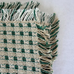 May include: A woven textile placemat featuring a grid pattern of small dark green squares on a natural beige background. The edges of the placemat are finished with a decorative fringe made of alternating beige and dark green thread strands.