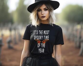May include: A woman wearing a black witch hat and a black t-shirt with the text "It's the most spooky time of the year" and a pair of orange eyes.