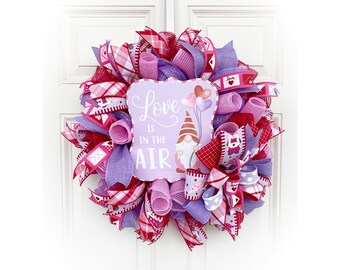 May include: A Valentine's Day wreath with pink, red, and purple mesh ribbon, a gnome, and a sign that says "Love is in the air".