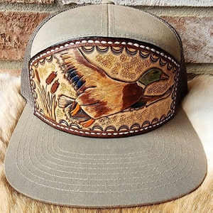 Puede incluir: Una gorra de camionero de color marrón claro y marrón con un parche de cuero que presenta una talla detallada de un pato silvestre en vuelo. El parche está cosido en la parte delantera de la gorra.