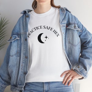 Puede incluir: Camiseta blanca con el texto "Practice Safe Hex" en letras negras, con un gráfico de luna creciente y estrellas. La camiseta la lleva puesta una persona que lleva una chaqueta vaquera.