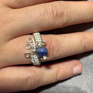 May include: A handmade silver wire ring with a blue gemstone bead. The ring has an intricate, openwork design with coiled and looped wire elements. The gemstone is a deep blue colour.