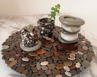 May include: A round stone coaster featuring a mix of brown, grey and white stones. Two small potted plants and a stack of stones with a candle on top are displayed on the coaster.