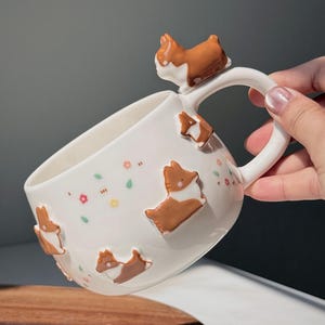 Può includere: Tazza in ceramica bianca decorata con figure di cani corgi marroni e bianchi. Il manico presenta una figura di corgi. La tazza è adornata da piccoli disegni floreali e di foglie in colori pastello. Un design affascinante e stravagante per gli amanti dei cani.