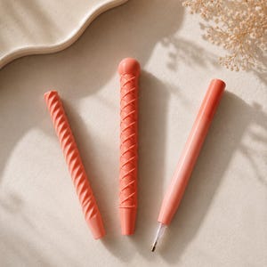 Può includere: Tre penne color corallo con texture distinte. Una penna presenta un motivo a spirale, un'altra ha un design a reticolo incrociato con una parte superiore arrotondata e la terza è una penna cilindrica liscia con una punta metallica visibile.