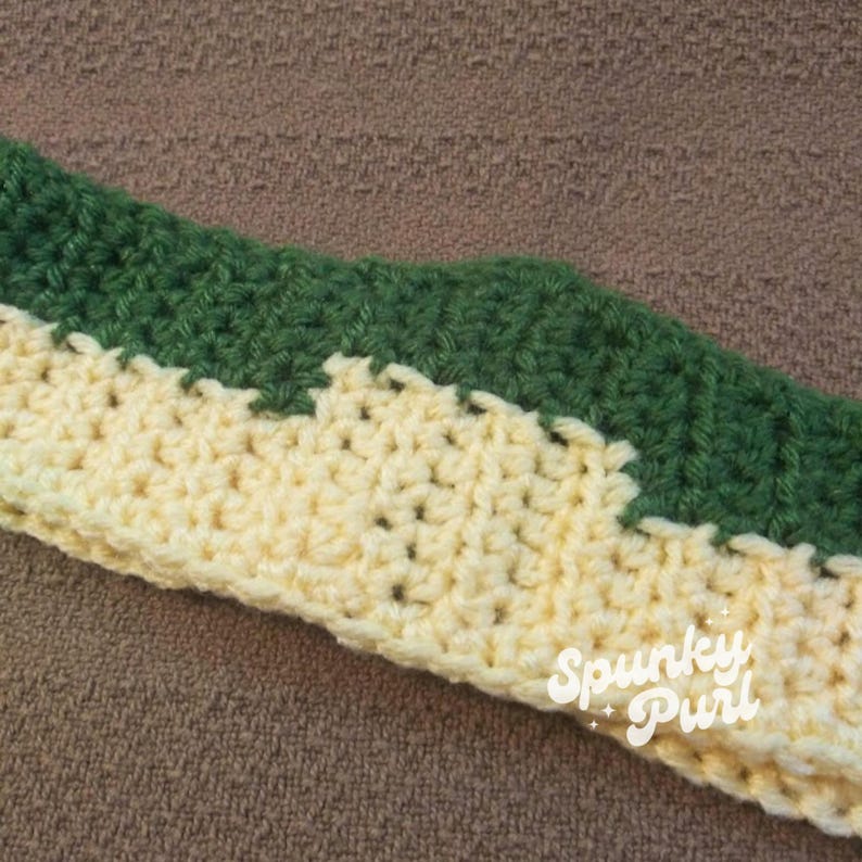 May include: A close-up of a crocheted headband with a yellow and green striped pattern. The headband is made with a soft, textured yarn and has a subtle sheen. The text "Spunky Purl" is visible on the headband.