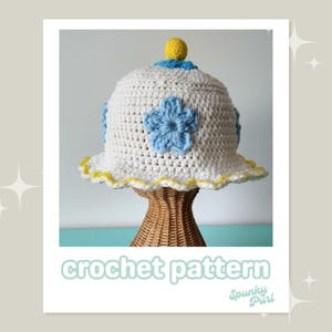 Op de afbeelding: Witte gehaakte emmerhoed met een gele pompon bovenop en blauwe bloemenaccenten. De hoed staat op een rieten hoedenstandaard. De tekst "crochet pattern" staat onder de hoed en "Spunky Purl" staat onder de tekst.