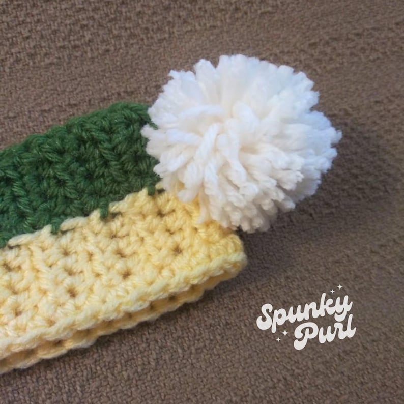 May include: A crocheted hat with a yellow and green striped brim and a white pom-pom on top. The hat is made with a soft yarn and has a comfortable fit. The hat is labeled "Spunky Purl".