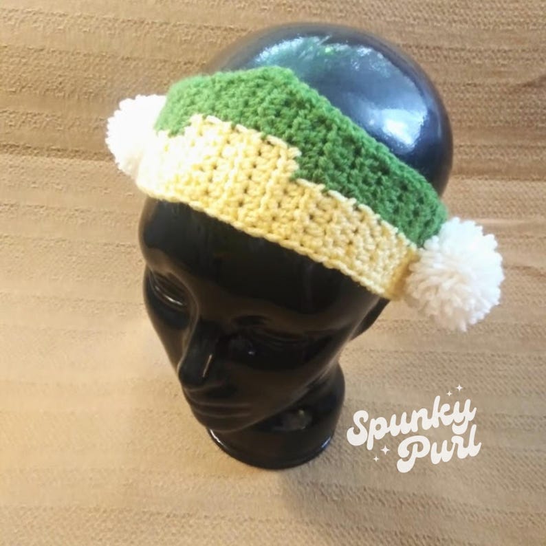 May include: A green, white, and yellow crocheted headband with two white pom-poms on a black mannequin head. The headband is made with a simple stitch pattern and has a striped design. The headband is a handmade item and is perfect for adding a touch of color to any outfit.