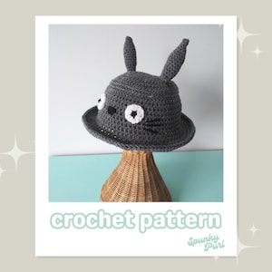 May include: A gray crochet hat with bunny ears, white eyes, and black whiskers. The hat is displayed on a wicker stand. The text "crochet pattern" is at the bottom of the image.