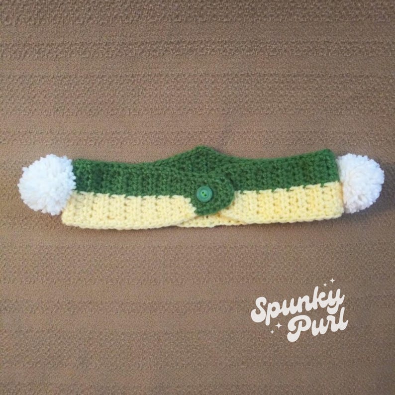 May include: A green and yellow crocheted dog collar with white pom-poms on each end and a green button in the center. The collar is on a brown surface. "Spunky Purl" is written in white on a black background in the lower right corner of the image.