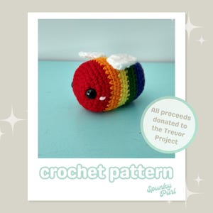 Puede incluir: Un patrón de crochet para una abeja arcoíris con cabeza roja y alas blancas. El patrón está disponible para su compra y todas las ganancias se donan al Proyecto Trevor. El texto "crochet pattern" y "Spunky Purl" es visible en la imagen.