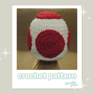 May include: A crocheted mushroom hat with a white base and red top. The hat has a large red circle on the top and two smaller red circles on the sides.