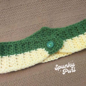 May include: A green and yellow crocheted headband with a green button. The headband is folded in half and the button is in the center of the fold.