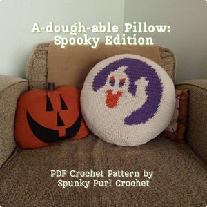 May include: A round white crochet cushion featuring a purple moon and a white ghost with orange eyes and mouth, next to an orange pumpkin-shaped cushion with black facial features. Text reads: A-dough-able Pillow: Spooky Edition PDF Crochet Pattern.