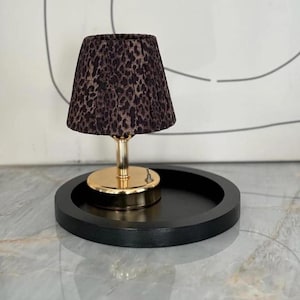 Può includere: Una piccola lampada da tavolo con paralume leopardato. La lampada ha una base e uno stelo color oro e poggia su un vassoio rotondo nero. La lampada è un elemento decorativo.