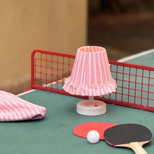 May include: A miniature ping pong table set with a red net, paddles, and a white ball. A small lamp with a pink and white striped shade sits on the table. A matching pink and white striped cloth is also present.